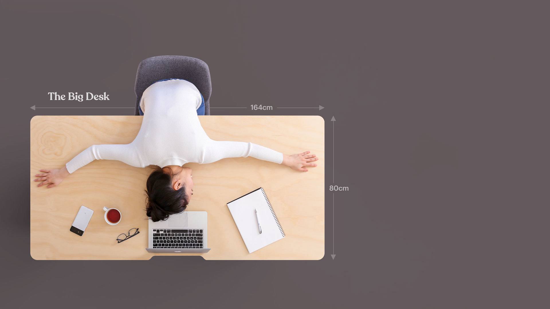 An overhead photo of a woman laying over and hugging her big desk, spreading out her arms.